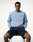 Man wearing a light blue sweatshirt and navy shorts sitting on a white stool against a white background