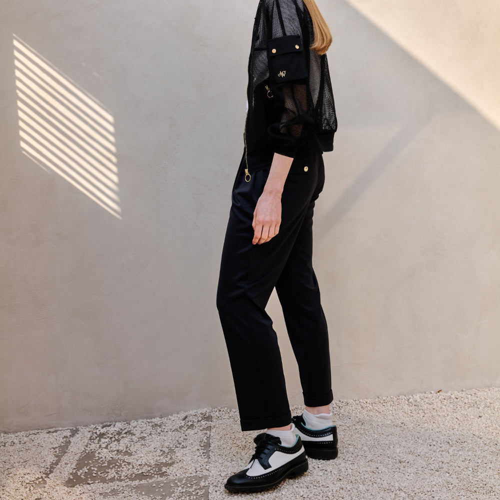 A model is shown from the side wearing a black sheer mesh long-sleeve top, black wide-leg trousers, white socks, and black-and-white platform sneakers, standing against a textured beige wall with diagonal shadows.