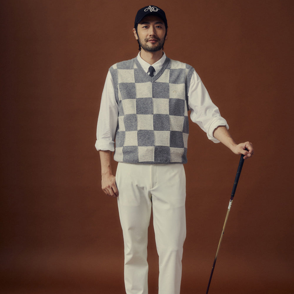 A man wearing a gray and white checkered sweater vest over a white long-sleeve shirt, white trousers, a black baseball cap, and holding a golf club, set against a solid brown studio background.