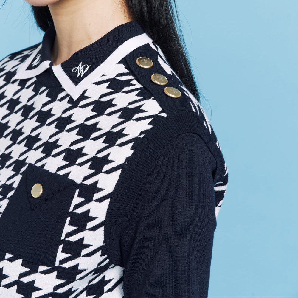 A close-up of a model wearing a black and white houndstooth patterned knit sweater vest with gold buttons, paired with a black cap, against a solid light blue background.