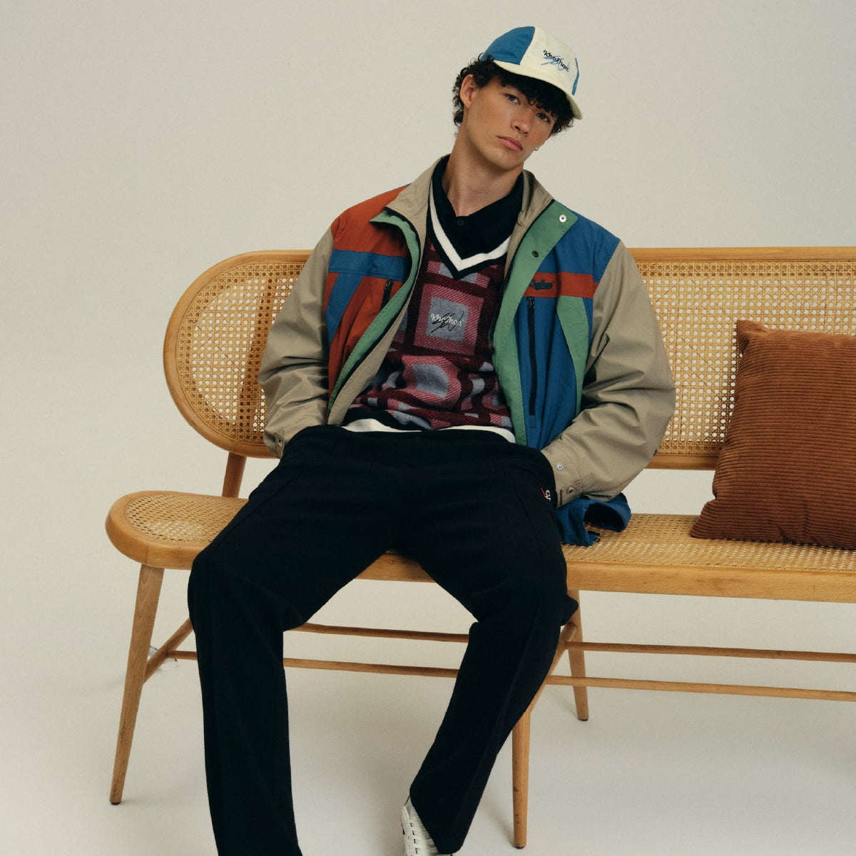 A model sits on a wooden cane bench wearing a multicolor retro colorblock jacket, black pants, and a blue and white cap against a plain off-white background.