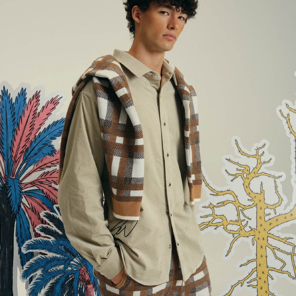 A model wearing a beige long-sleeve button-up shirt with a black embroidered signature on the chest, paired with brown and beige plaid pants and a matching plaid hooded sweatshirt. The background is a white wall decorated with colorful tropical and tree illustrations.
