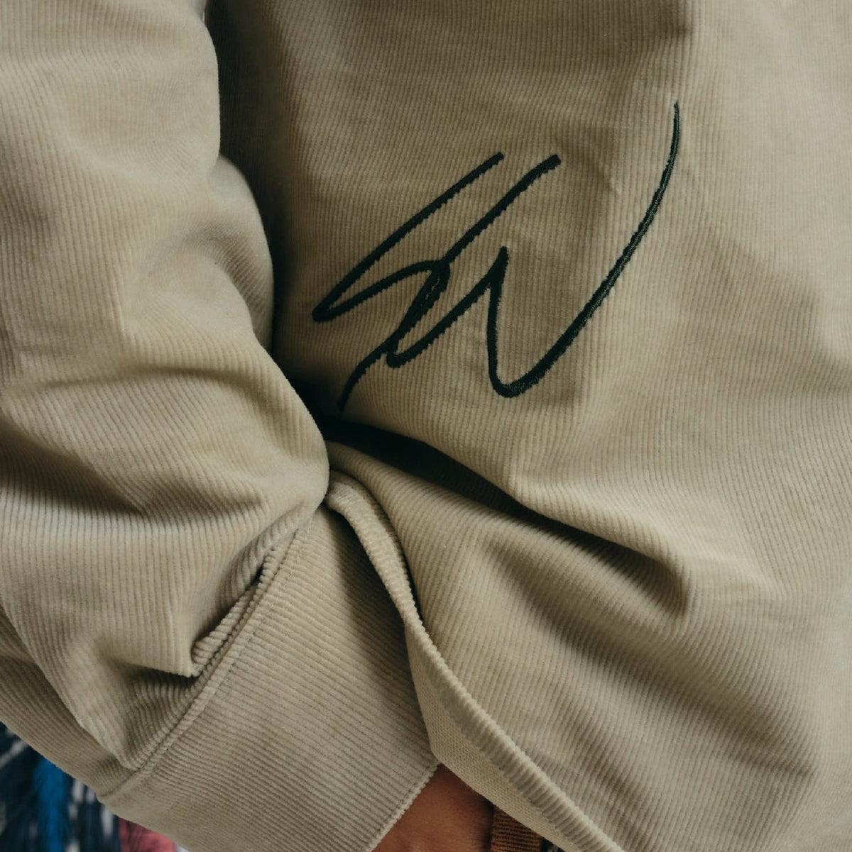 A model wearing a beige long-sleeve button-up shirt with a black embroidered signature on the chest, paired with brown and beige plaid pants and a matching plaid hooded sweatshirt. The background is a white wall decorated with colorful tropical and tree illustrations.