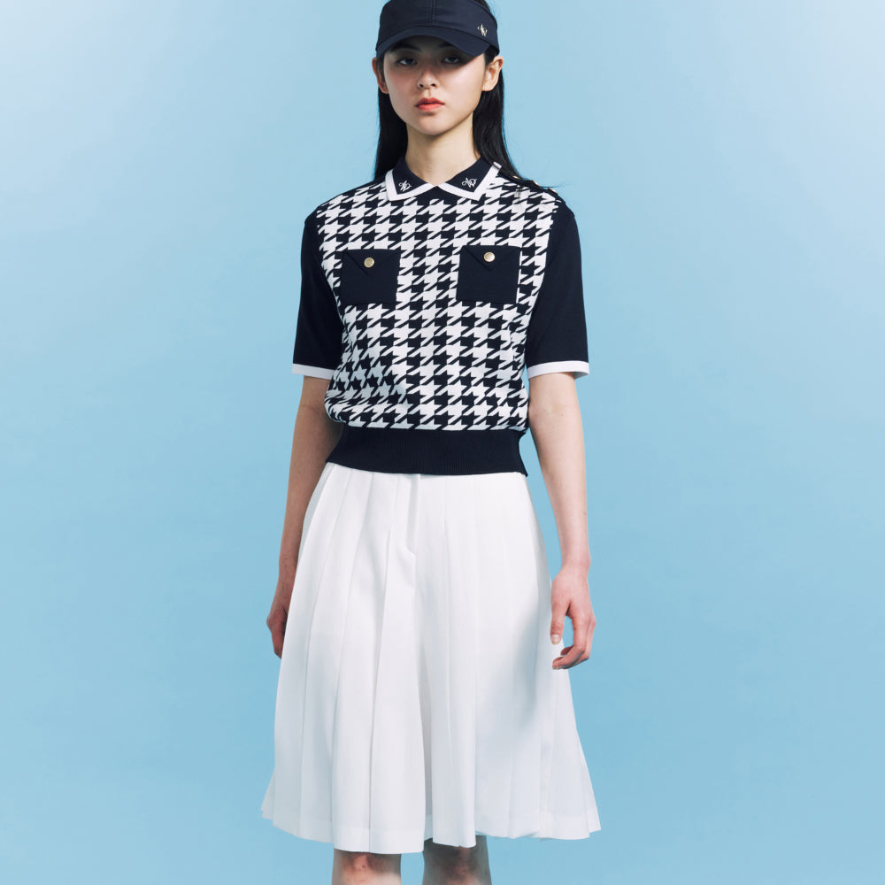A model stands against a light blue studio background wearing a black and white houndstooth short-sleeve knit polo shirt, a white pleated midi skirt, a black baseball cap, white socks, and black shoes.