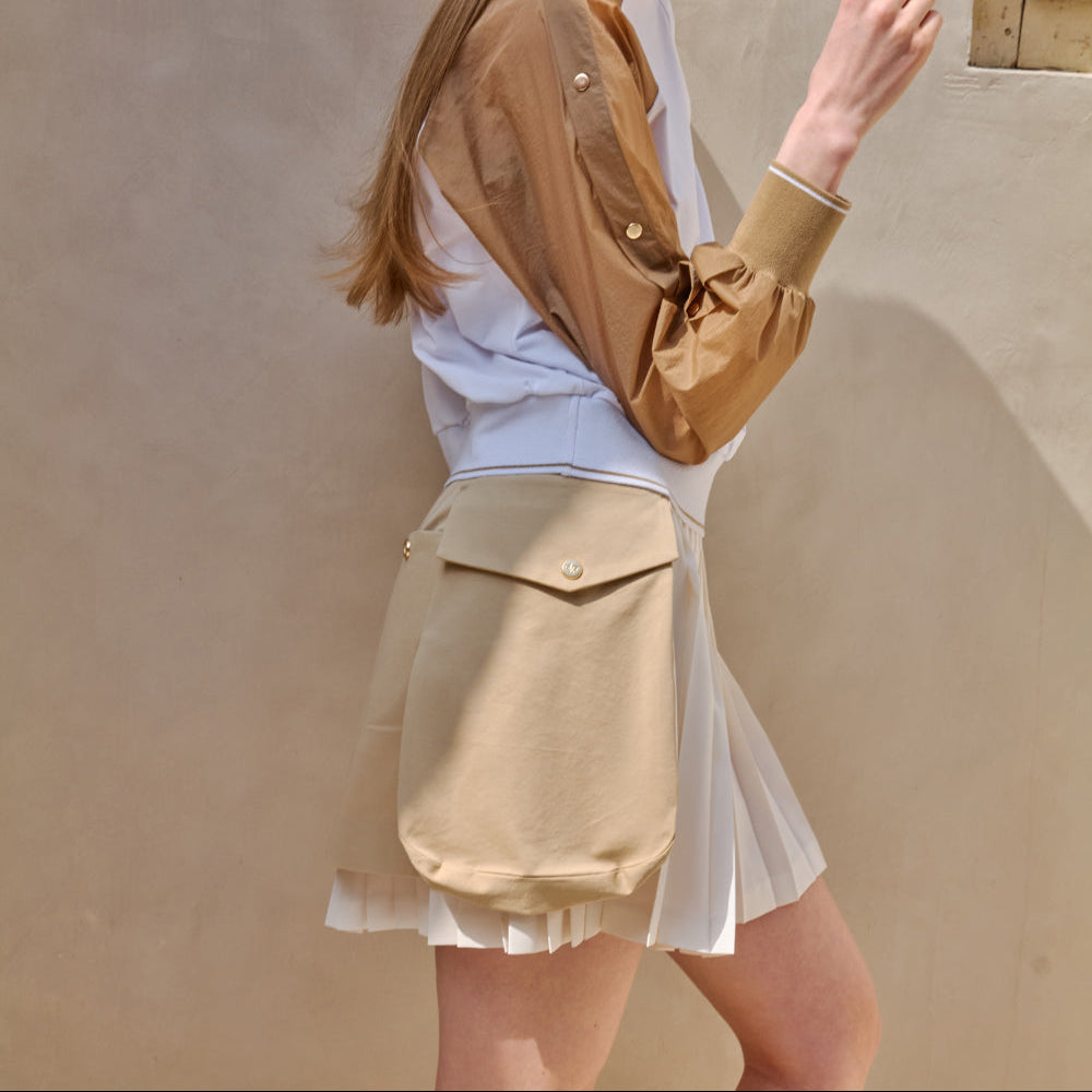 A model wearing a beige utility-style pleated mini skirt with a large front flap pocket, styled over a white top and a tan button-up overshirt, set against a textured beige wall.