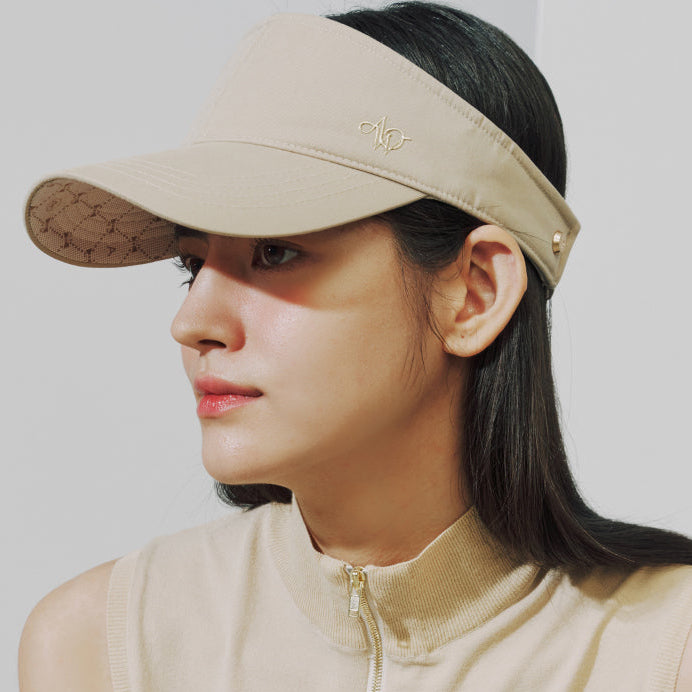A woman wearing a beige visor hat with a small gold monogram logo on the front, paired with a matching beige ribbed top. The visor has a subtle tonal patterned brim and a small gold rivet detail.