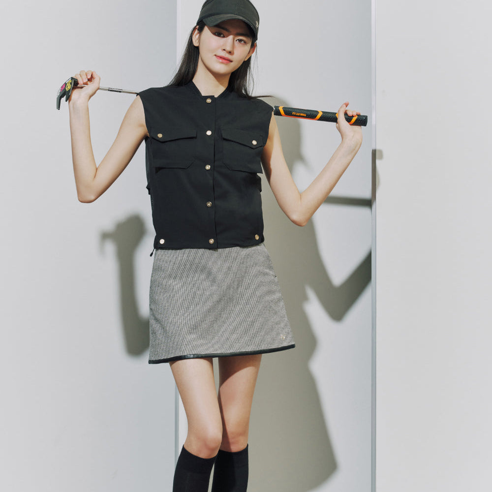 A woman stands against a white wall holding a baseball bat, wearing a black sleeveless vest with snap buttons, a gray textured mini skirt, black knee-high socks, and a dark baseball cap.