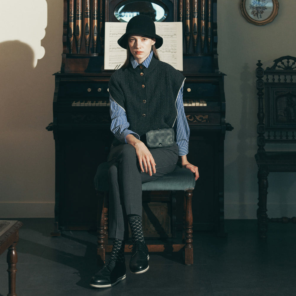 A person wearing a black bucket hat, black vest, blue striped shirt, and dark trousers sits on a wooden stool in front of a vintage dark wood organ or piano in a room with framed wall art.