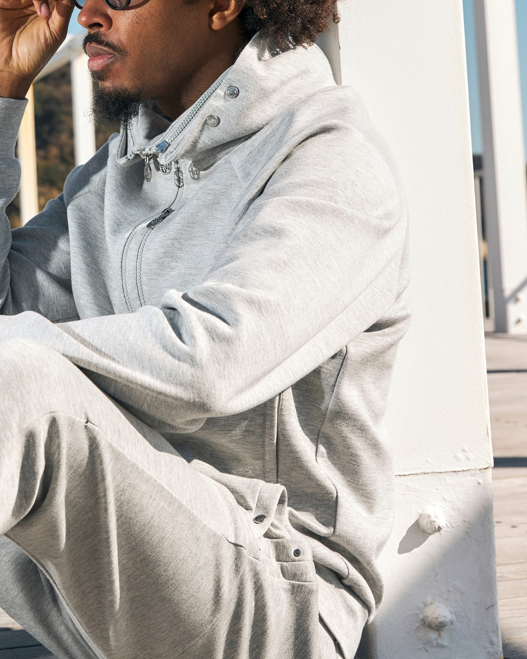 A man wearing a light gray heathered zip-up hoodie and matching sweatpants, sitting outdoors next to a white structural column.