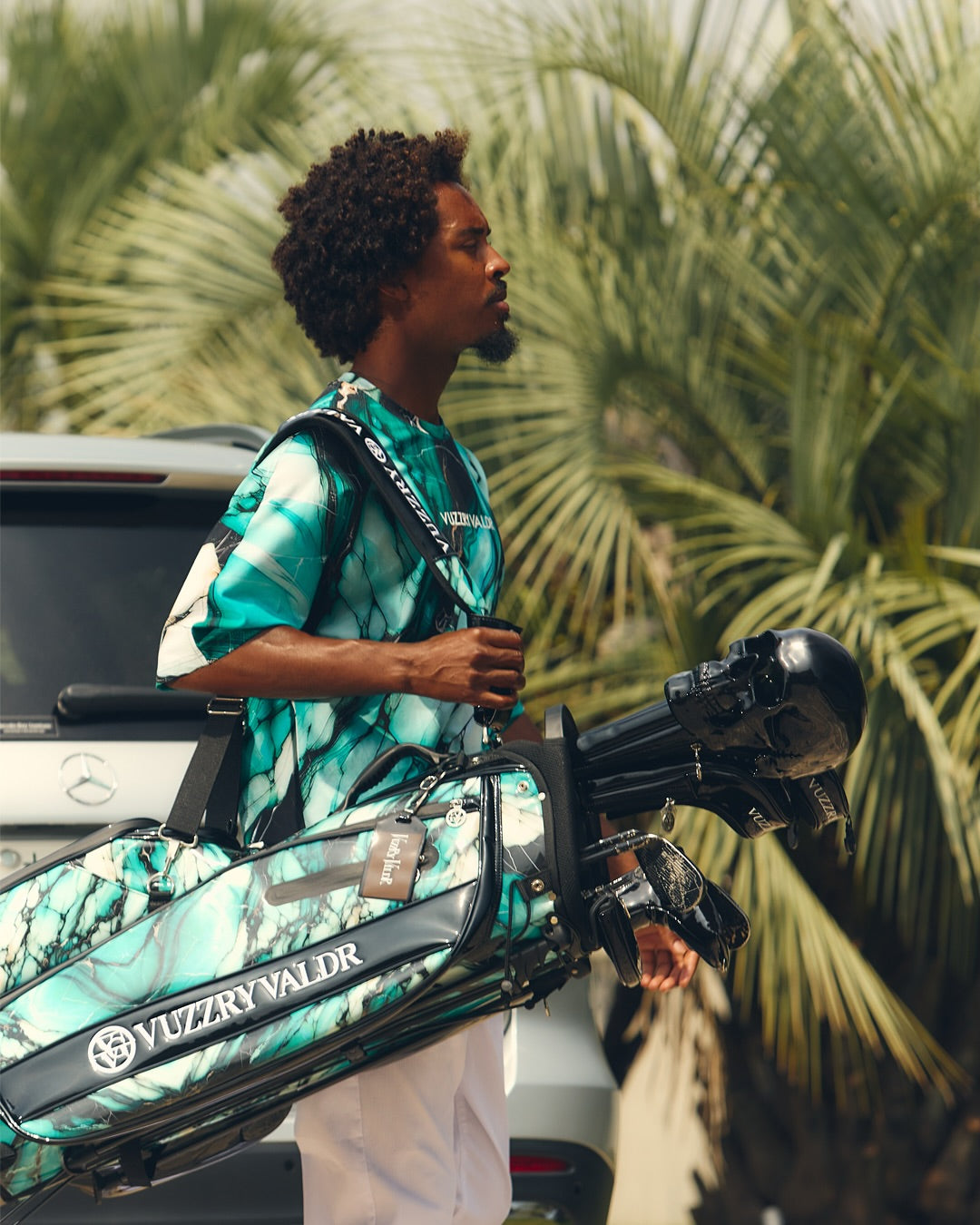 A man wearing a teal and green tie-dye Vizzary Valdr golf shirt and white pants carries a matching Vizzary Valdr golf bag with clubs, set against a background of palm trees and a white vehicle.