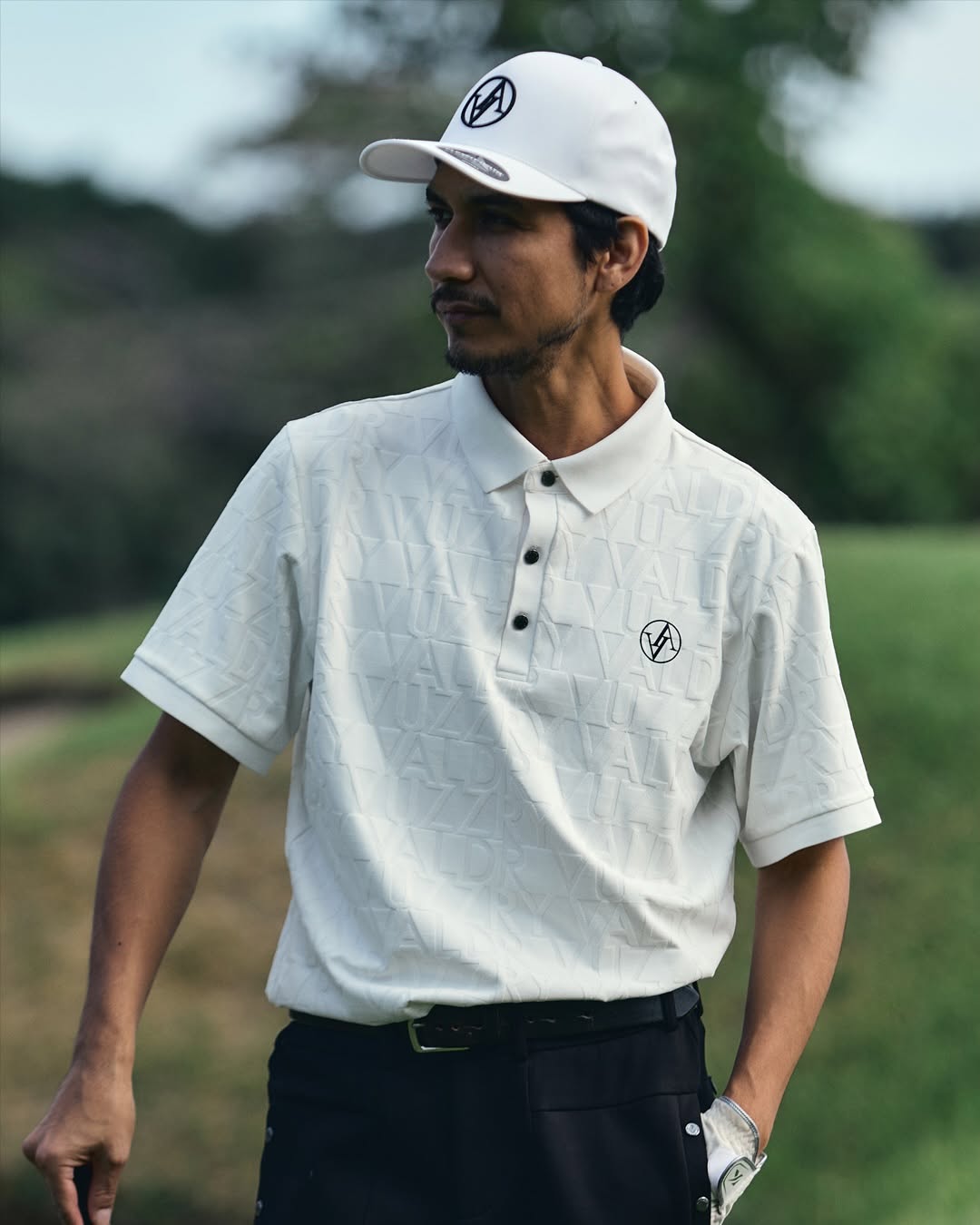 A man wearing a white textured short-sleeve polo shirt and a white baseball cap with a circular logo, standing outdoors on a golf course.