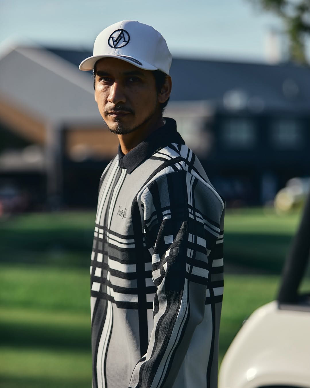 A man wearing a white baseball cap with a black circular logo and a gray and black striped long-sleeve shirt, standing outdoors on a grassy field with a blurred background.