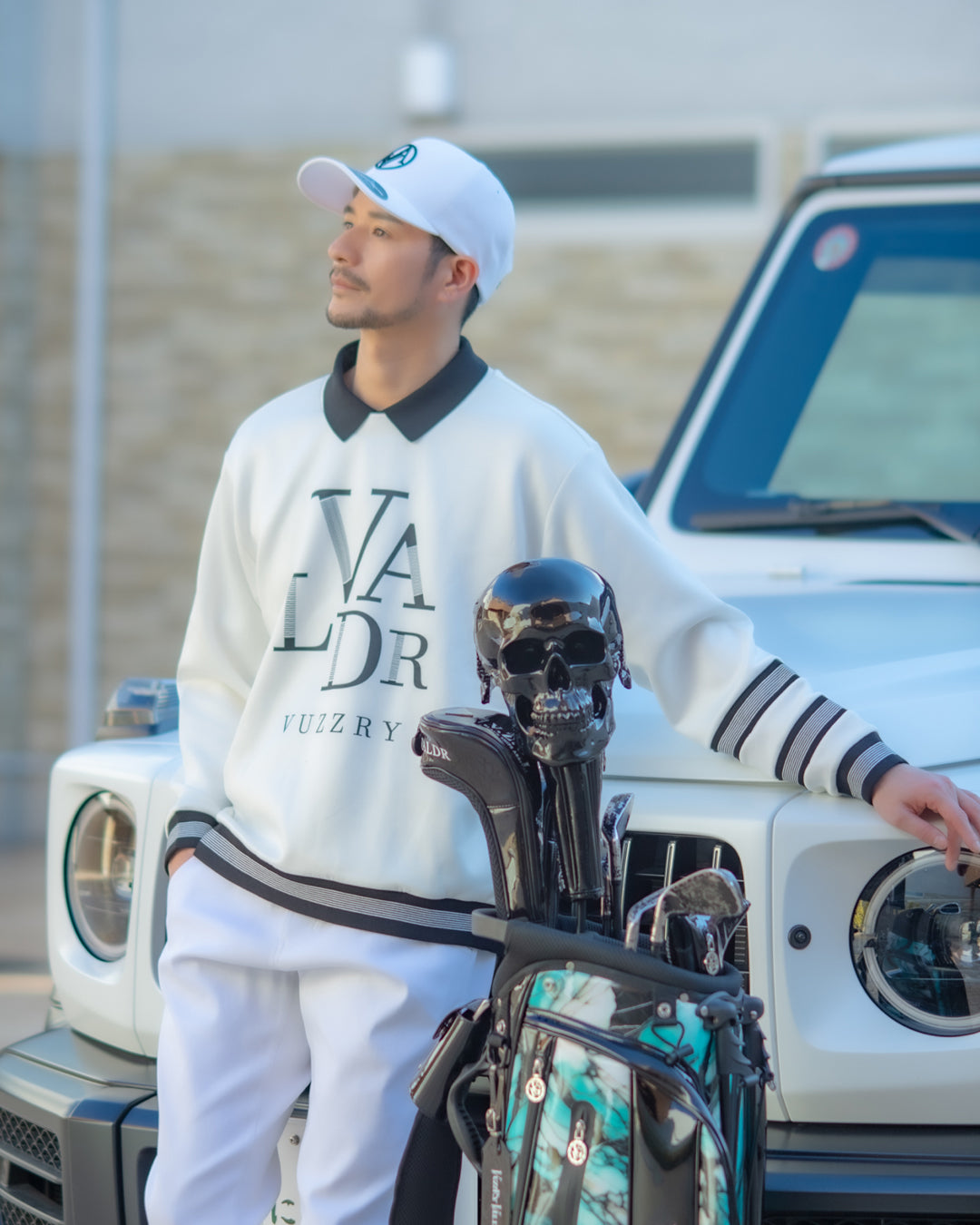 A man wearing a white and black pullover sweatshirt with 'NADLR' and 'VUZZRY' printed on the front, white pants, and a white cap, posing next to a white vehicle with a golf bag in the foreground.