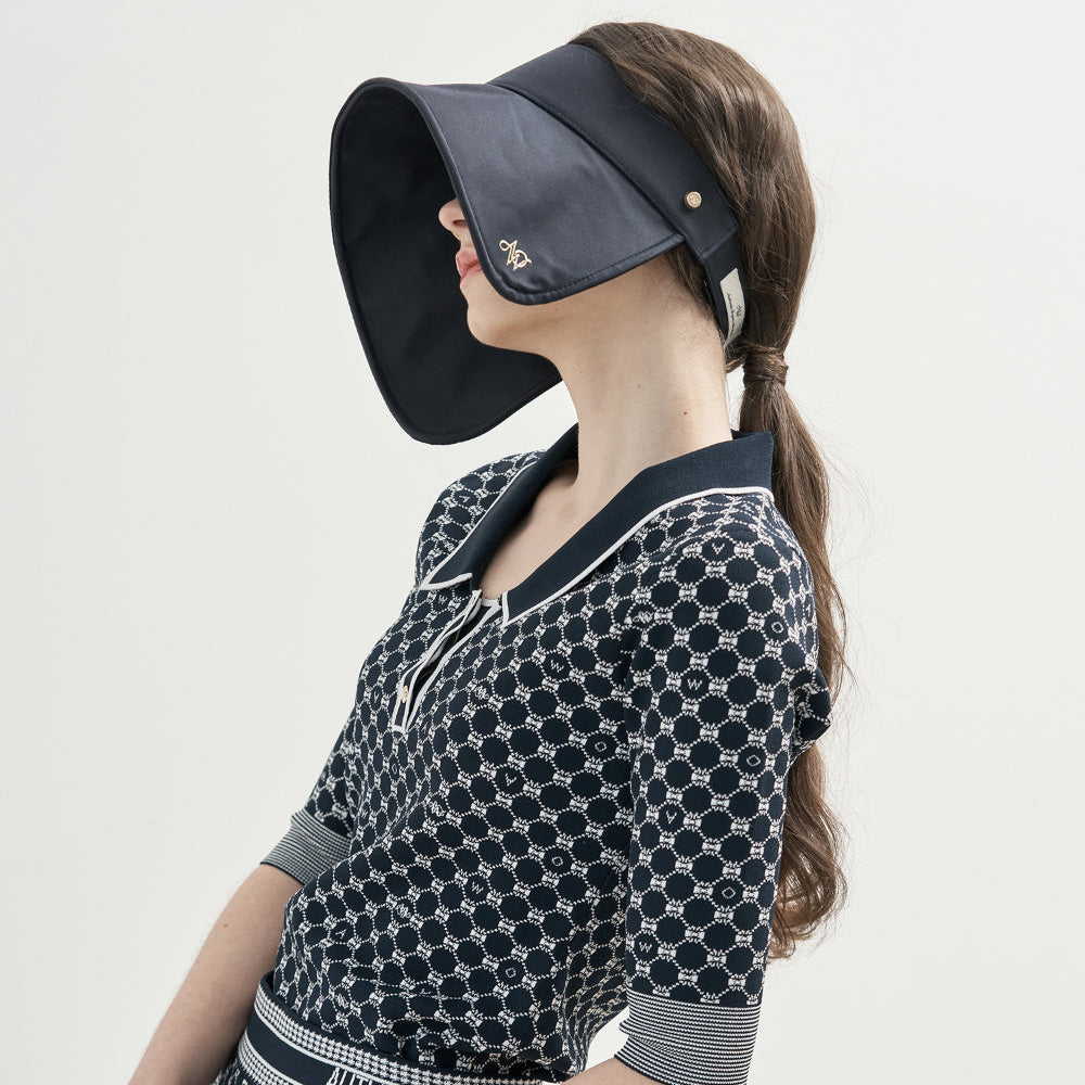A woman in profile wearing a dark navy sun visor and a matching navy and white patterned knit dress with a tie neckline, short sleeves, and a decorative waistband, photographed against a plain light background.