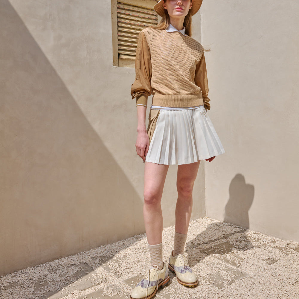 A model wearing a tan knit long-sleeve sweater, a white pleated mini skirt, beige socks, and cream lace-up shoes, standing in a sunlit corner with a beige stucco wall.