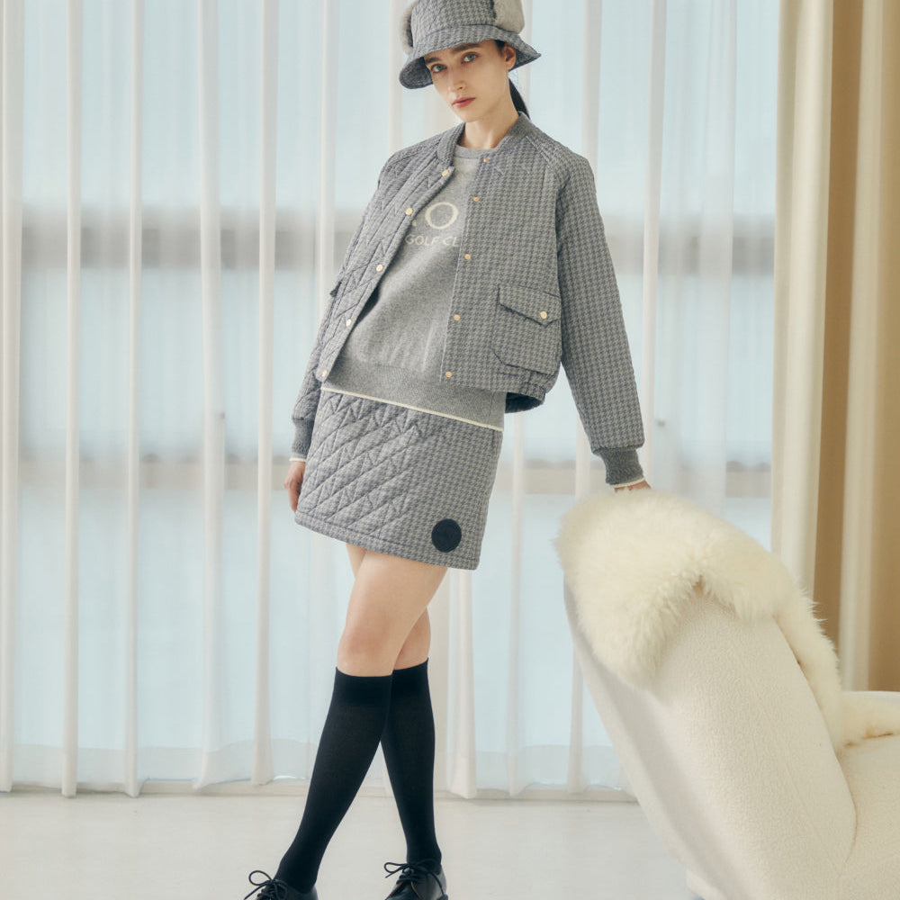 A model stands in a bright room wearing a gray and white checkered jacket, matching quilted mini skirt, a gray and white checkered bucket hat, black knee-high socks, and black shoes. To the right is a white armchair with a fluffy cream throw blanket.