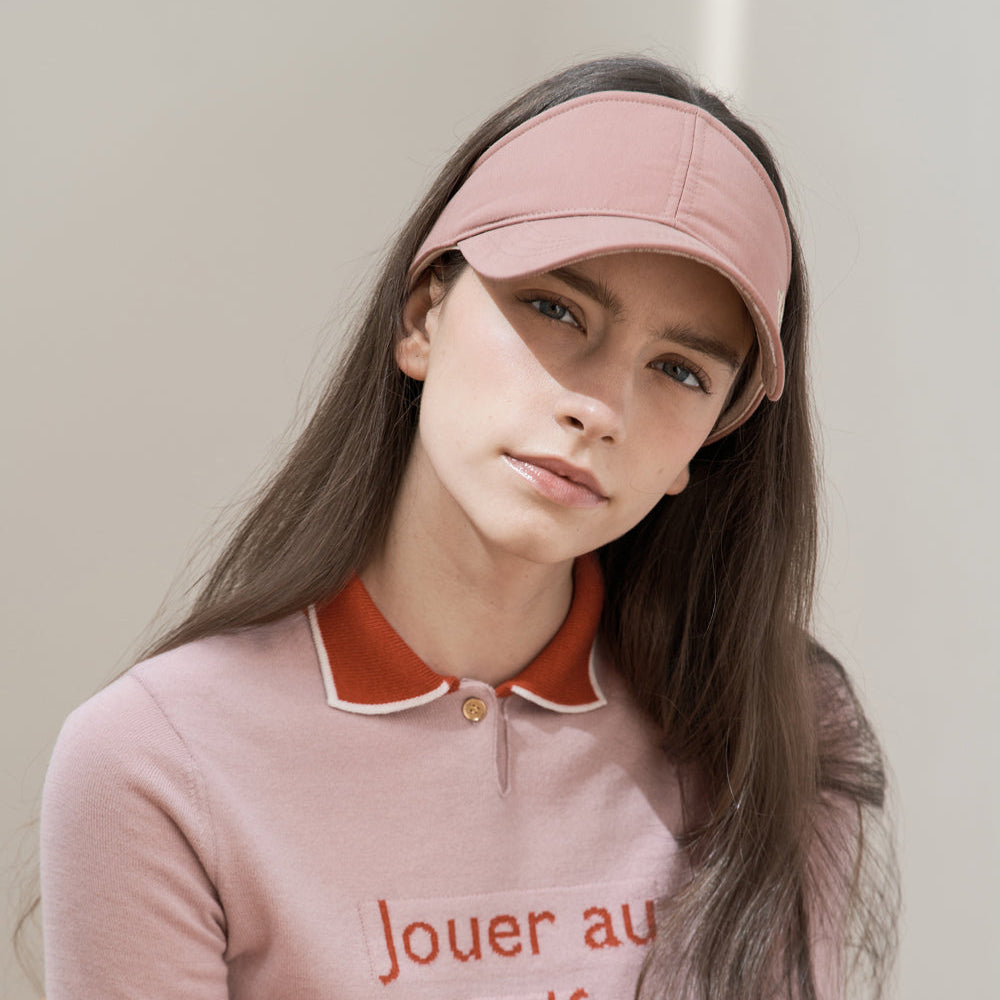 A woman wearing a dusty pink baseball visor and a matching pink long-sleeve knit sweater with a contrasting orange collar and orange text reading 'Jouer au golf' and 'bien'.