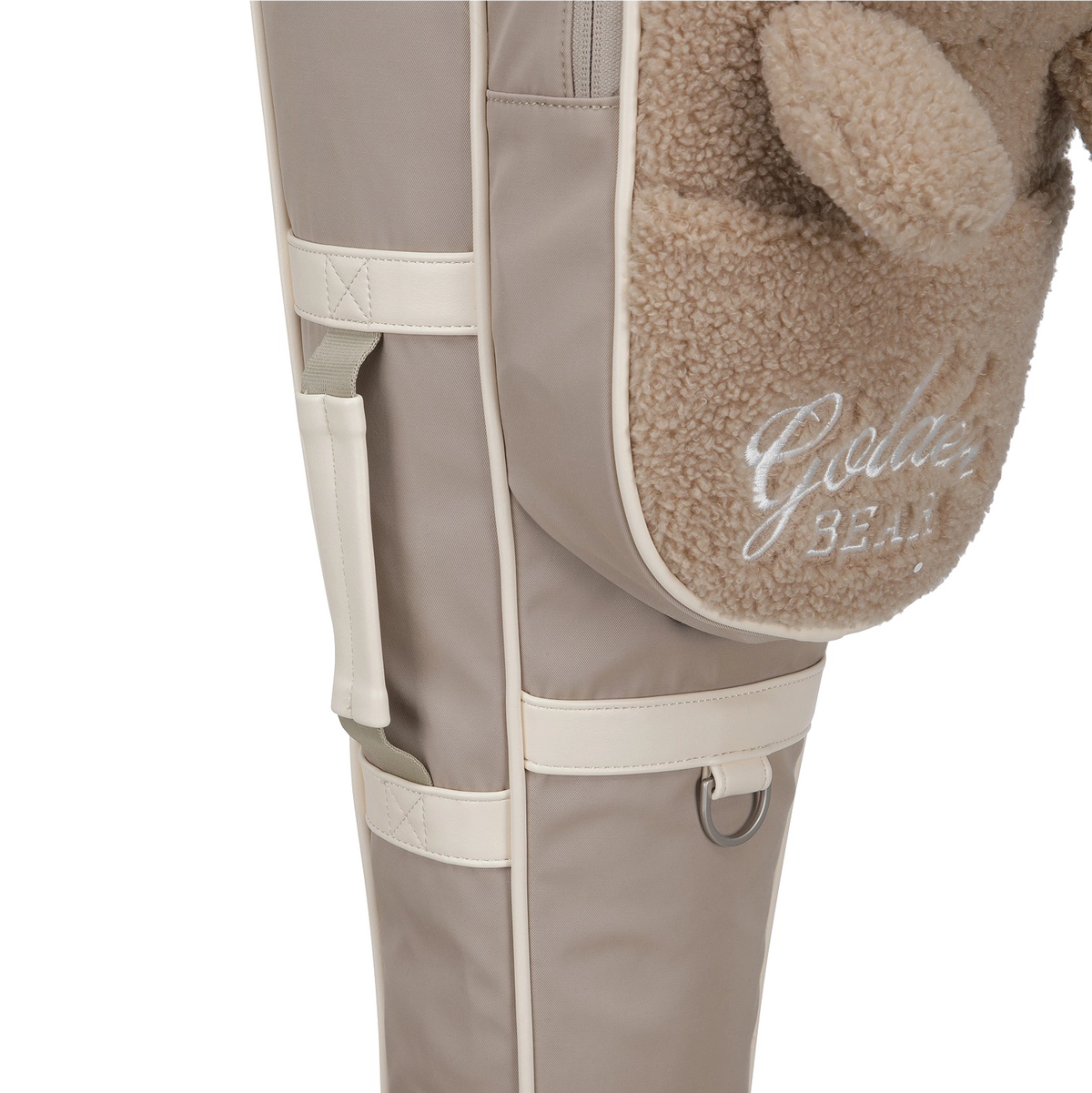 Close-up of a beige and cream padded bag with a plush brown teddy bear ear and pocket, featuring the embroidered text 'Golden Bear'.
