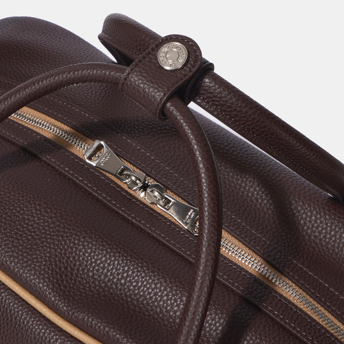 Close-up of a dark brown pebbled leather bag with a silver-tone zipper pull and a matching leather strap detail on a light gray background.