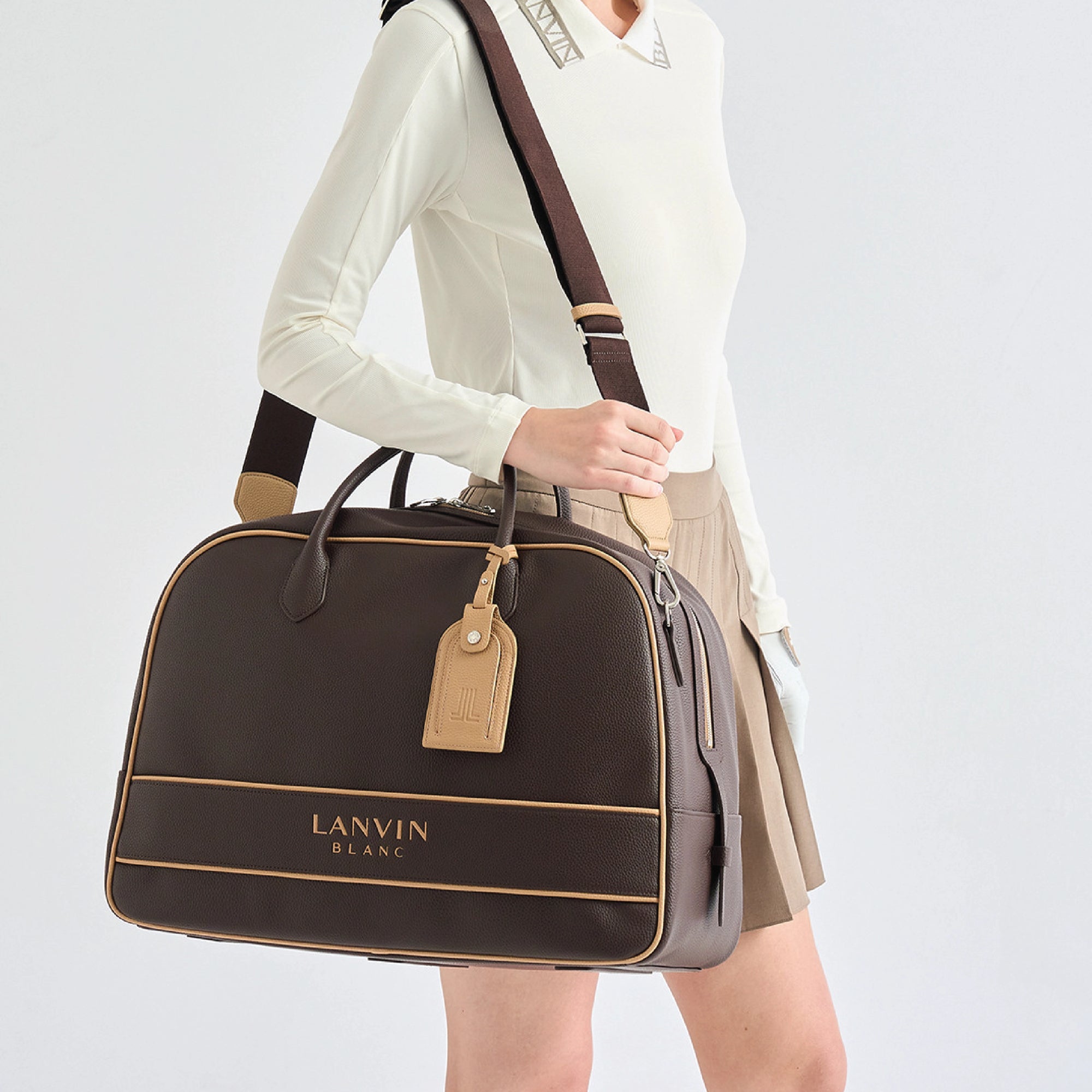 A model wearing a cream long-sleeve top and beige skirt carries a dark brown Lanvin Blanc duffle bag with tan trim and a matching leather luggage tag against a light gray studio background.