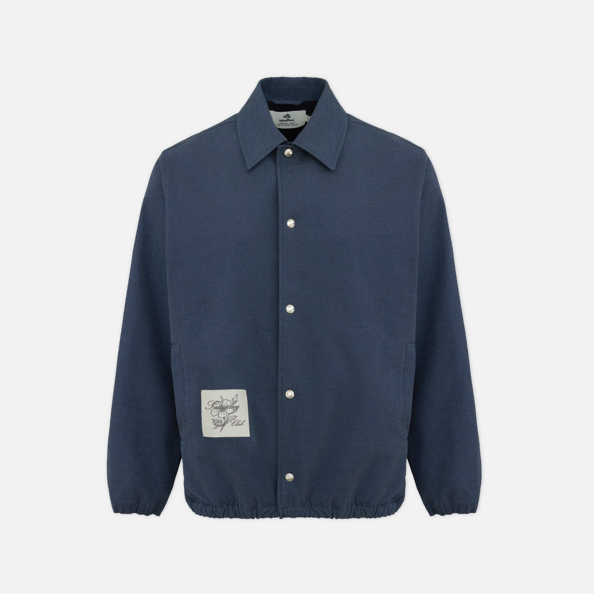 A navy blue cotton coach jacket with a pointed collar, snap-button front, and a square embroidered patch on the lower left front, shown against a light gray background.