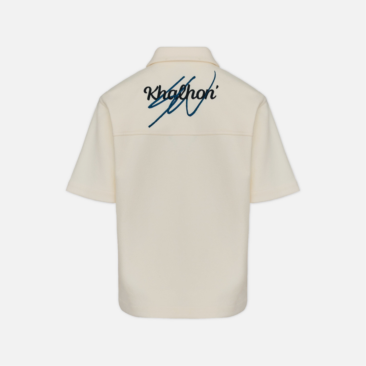 Back view of a cream-colored short-sleeve button-up shirt with a dark blue embroidered Khaiton logo on the upper back, displayed on a light gray background.