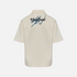 Back view of a cream-colored short-sleeve button-up shirt with a dark blue embroidered Khaiton logo on the upper back, displayed on a light gray background.