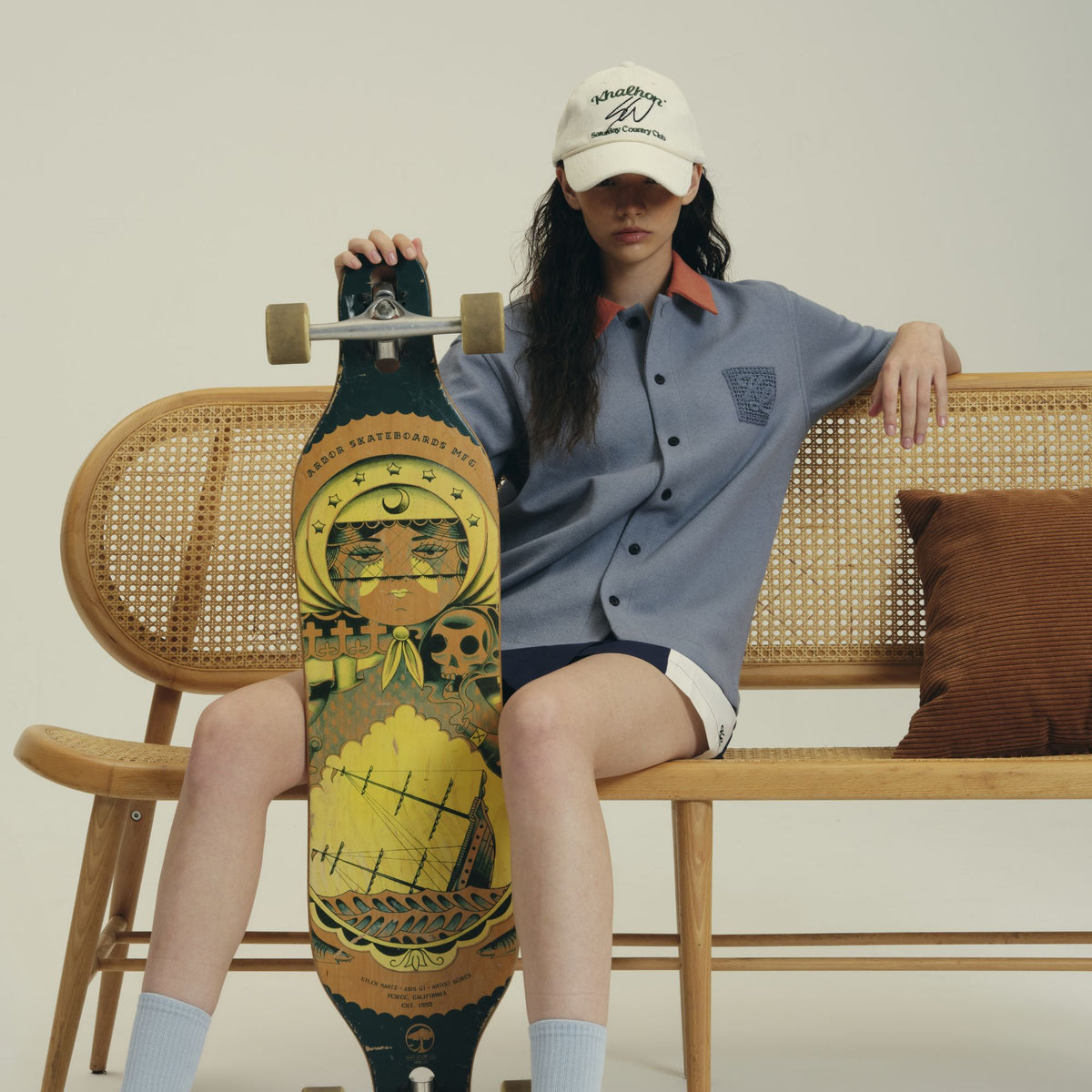 A model sits on a wooden cane bench holding a longboard skateboard with a detailed graphic design. The model is wearing a blue-gray button-up shirt, dark shorts, light blue socks, and a white baseball cap.