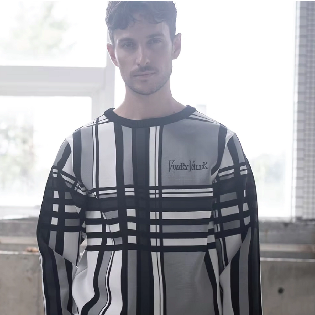 A man wearing a long-sleeve crewneck sweatshirt with a black, white, and gray abstract plaid and stripe pattern, featuring the brand name 'VUZZRY VALDR' printed on the chest.
