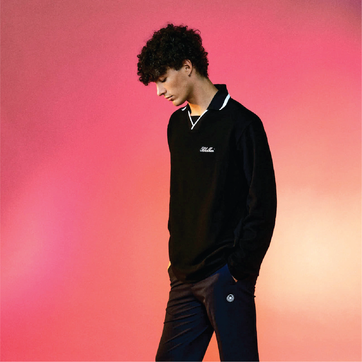 A man wearing a black long-sleeve polo shirt with white trim on the collar and a small white embroidered logo on the left chest, paired with dark pants, posed against a vibrant pink and orange gradient background.