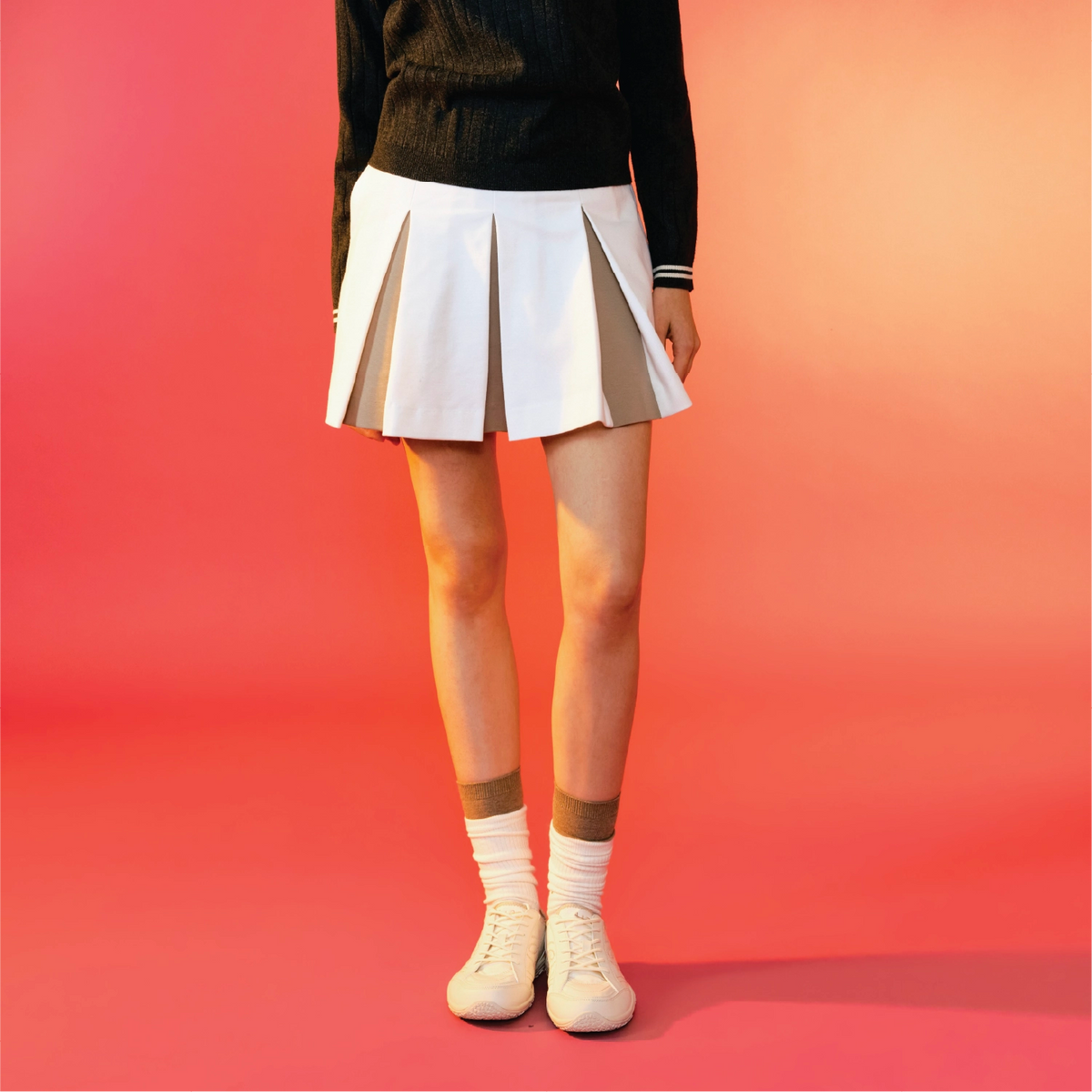 A model from the waist down wears a white pleated mini skirt with beige side panels, a black long-sleeve top, white socks, and white sneakers, set against a vibrant pink and orange gradient background.