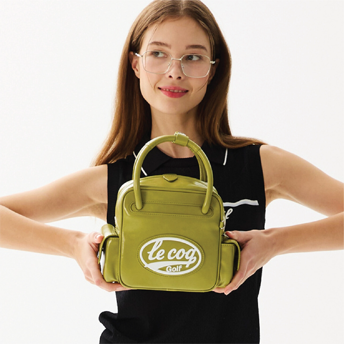 A woman wearing glasses and a black sleeveless top holds a small olive green handbag with top handles and side pockets. The front of the bag has a white oval logo reading 'le cog Golf'.
