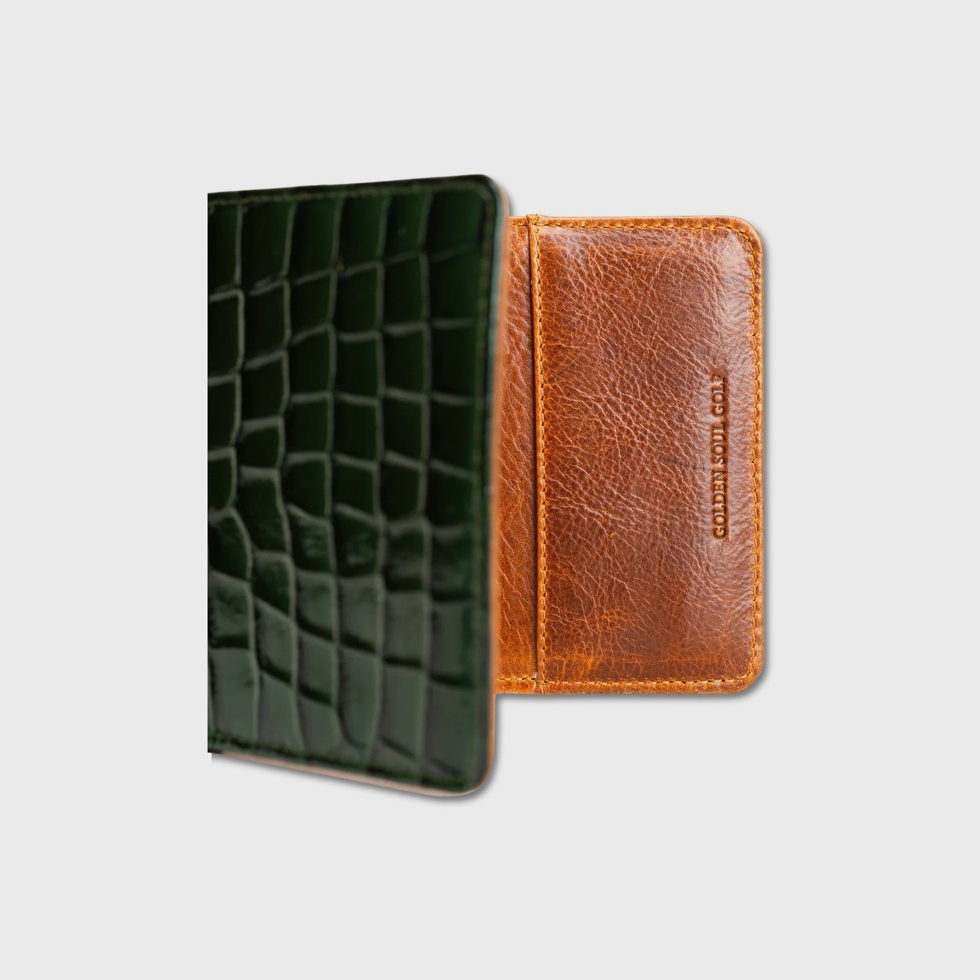 A yardage book holder shown side by side on a light gray background: one dark green with an embossed crocodile pattern, and one tan/cognac with visible grain and vertical debossed branding.