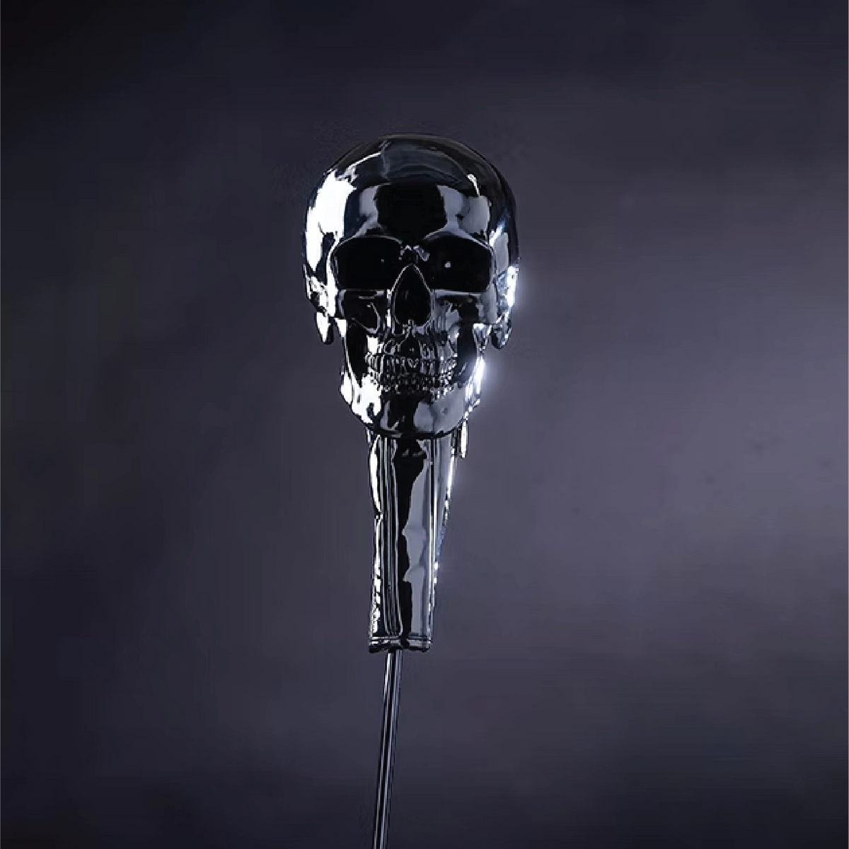 A polished chrome skull-shaped car key fob or decorative key fob with a reflective metallic finish, standing upright against a dark smoky gray background.