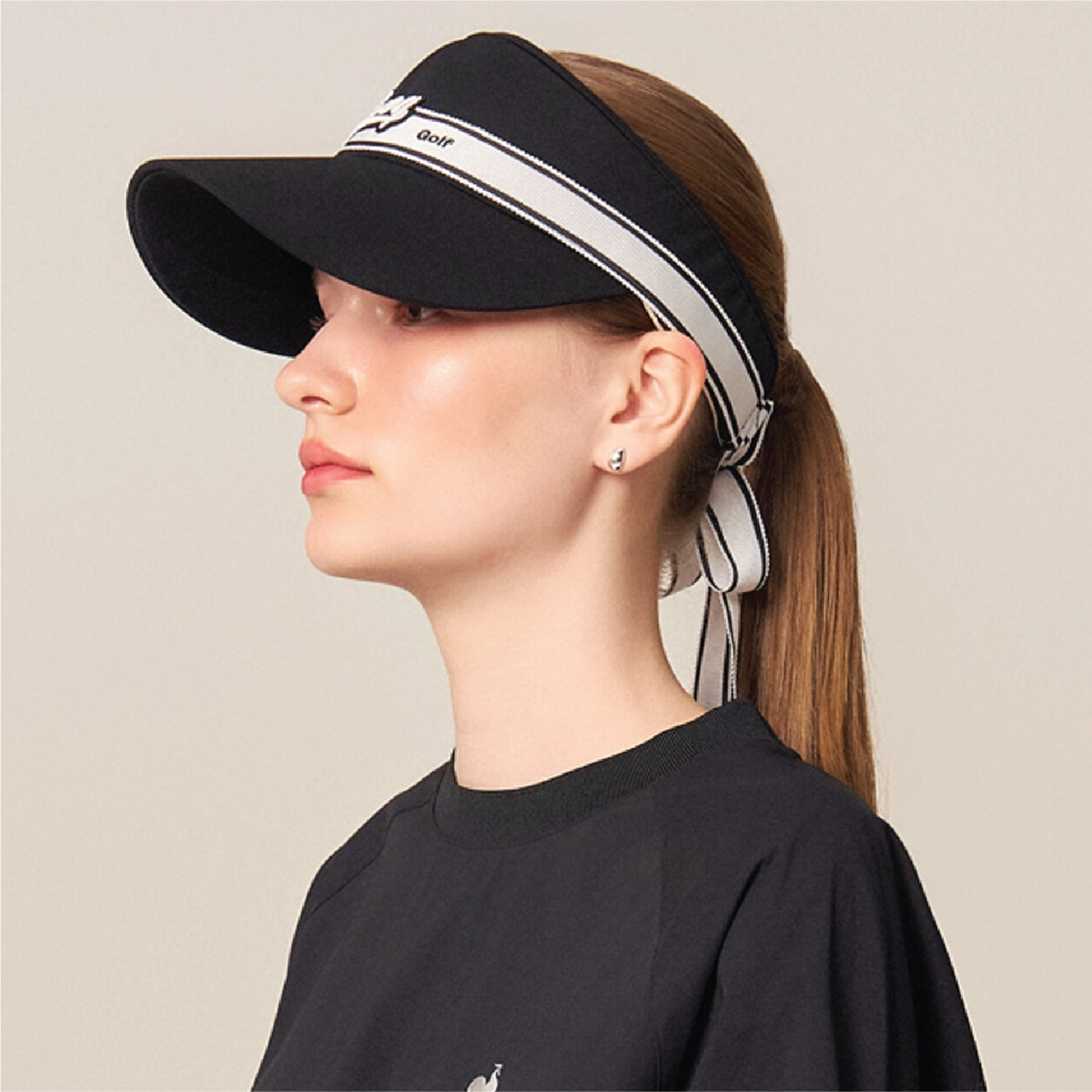 A woman in profile wearing a black visor with a white and gray striped headband detail and a matching tie-back ribbon, paired with a black crewneck sweatshirt against a plain beige background.