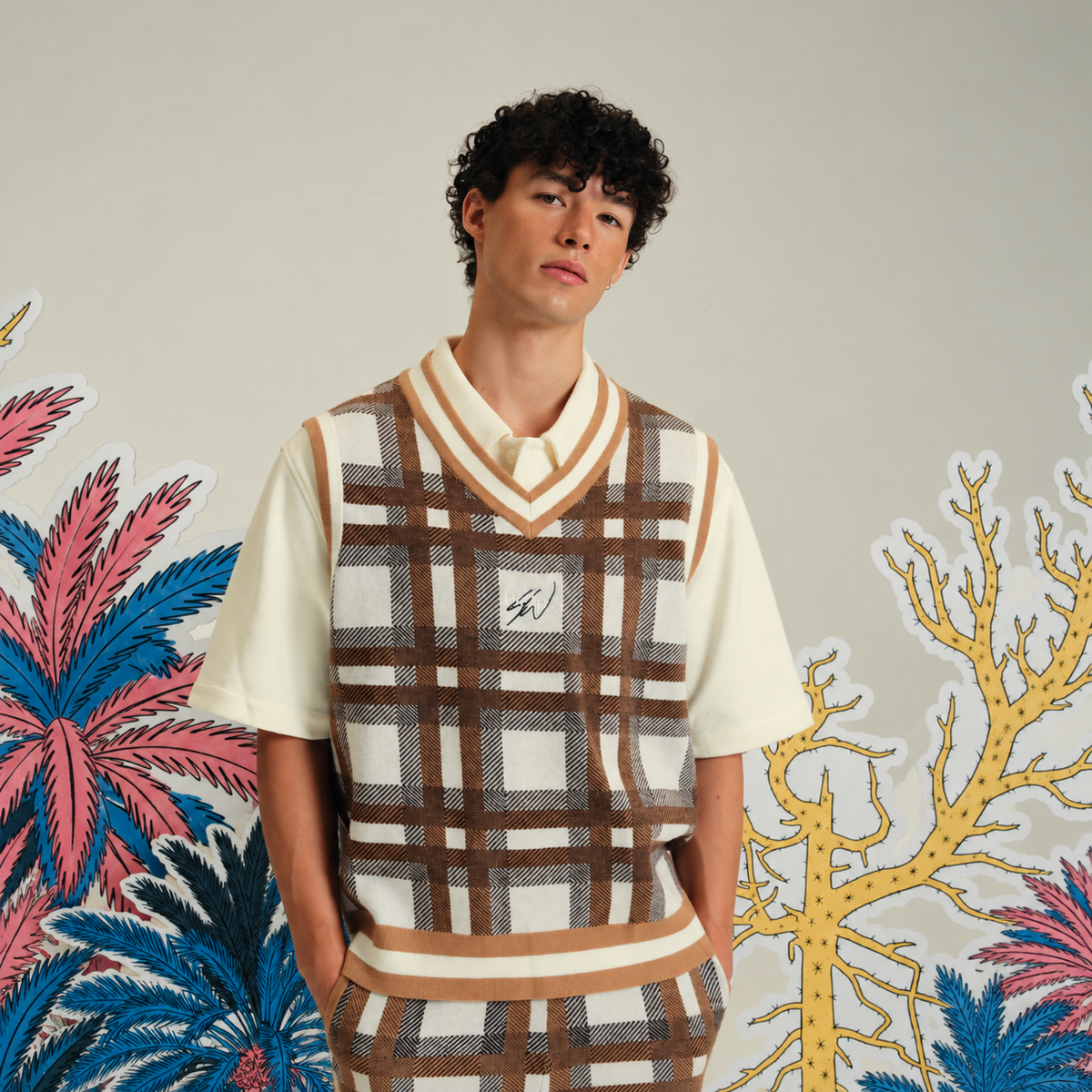A man wearing a cream short-sleeve knit polo shirt with a brown and beige plaid sweater vest layered over it, posed against a light gray wall with colorful tropical cutout decorations.