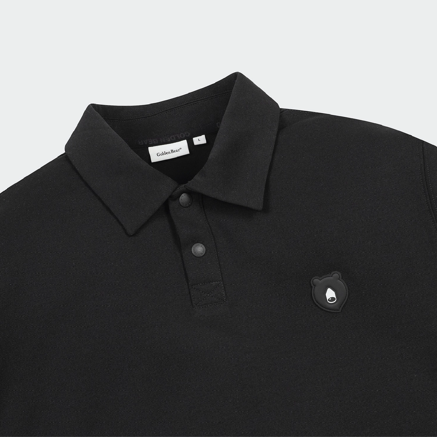 Close-up of a black polo shirt with a collar, two-button placket, and a small bear-shaped chest emblem. A neck label reads 'Golden Bear'.