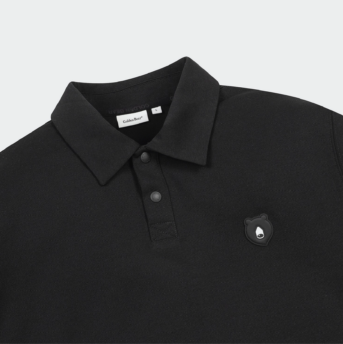 Close-up of a black polo shirt with a collar, two-button placket, and a small bear-shaped chest emblem. A neck label reads 'Golden Bear'.