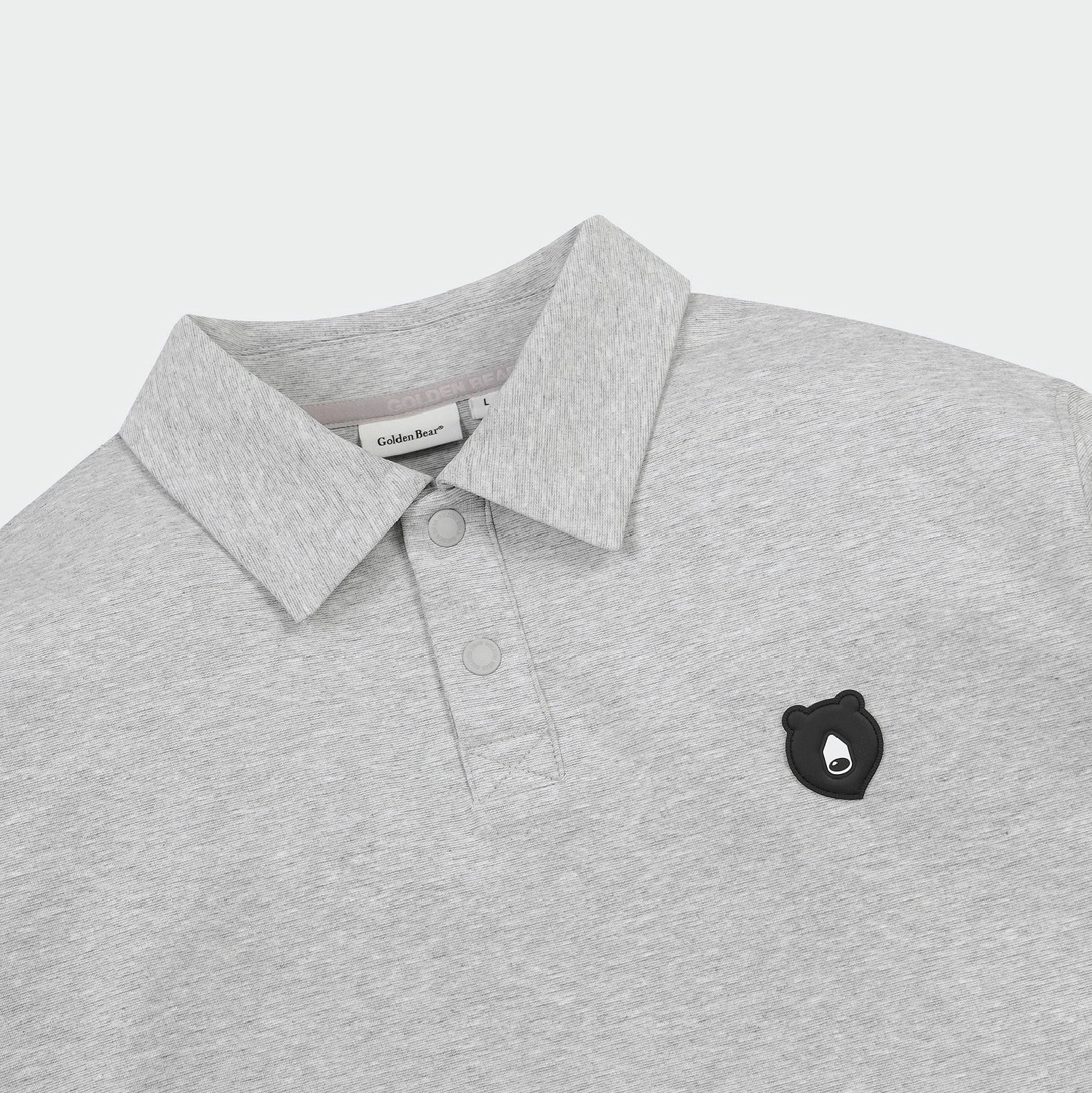 A close-up of a light gray heathered polo shirt by Golden Bear, featuring a classic collar, two-button placket, and a small black embroidered bear head logo on the left chest.