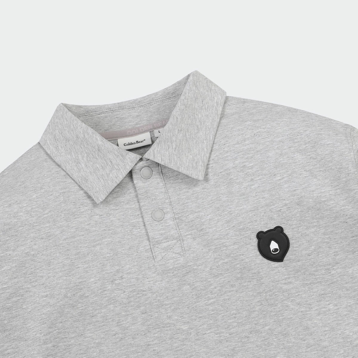 A close-up of a light gray heathered polo shirt by Golden Bear, featuring a classic collar, two-button placket, and a small black embroidered bear head logo on the left chest.