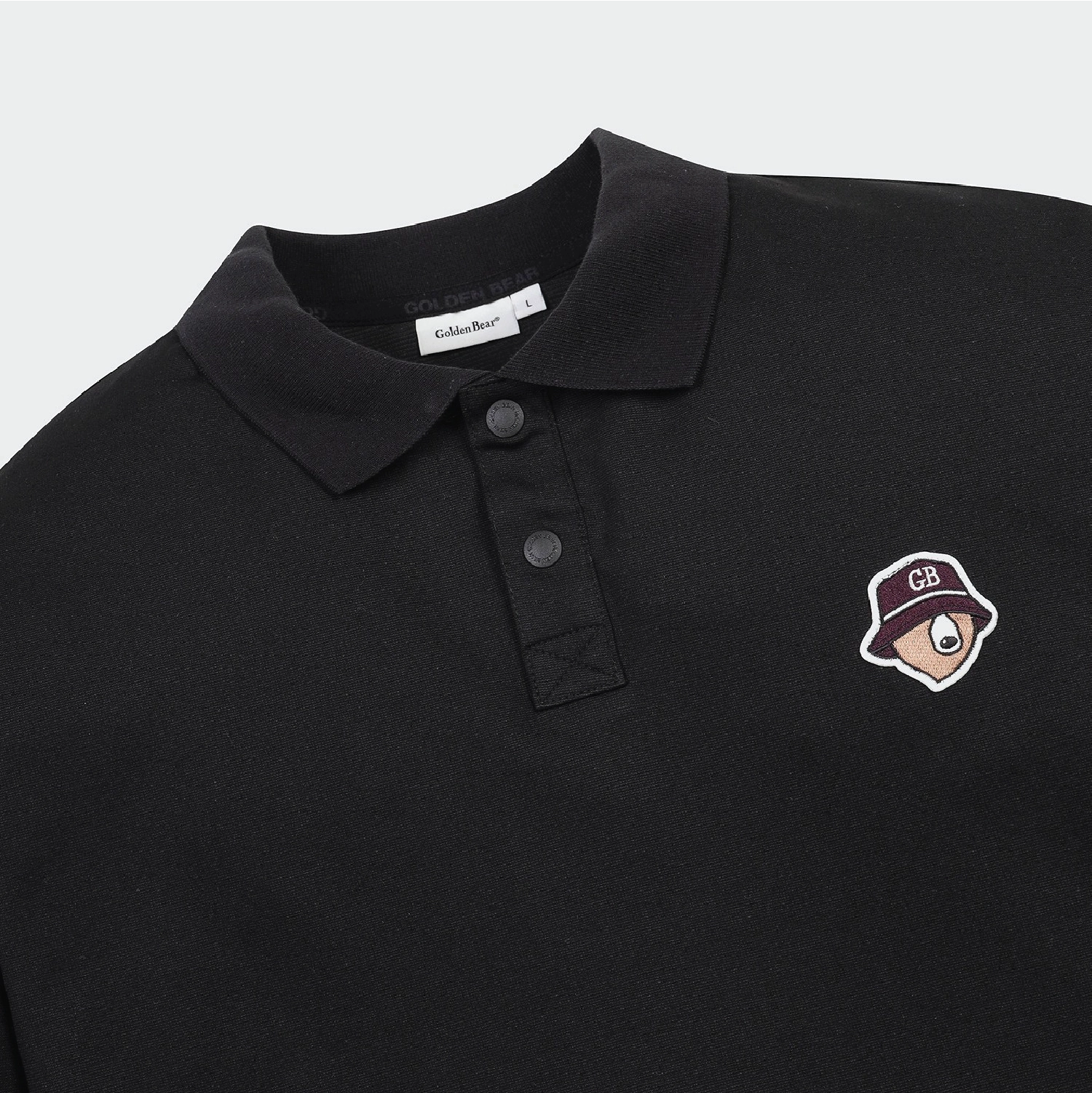 Close-up of a black polo shirt with a collar, three-button placket, and a small embroidered cartoon hat patch on the left chest. The neck label reads Schuber.