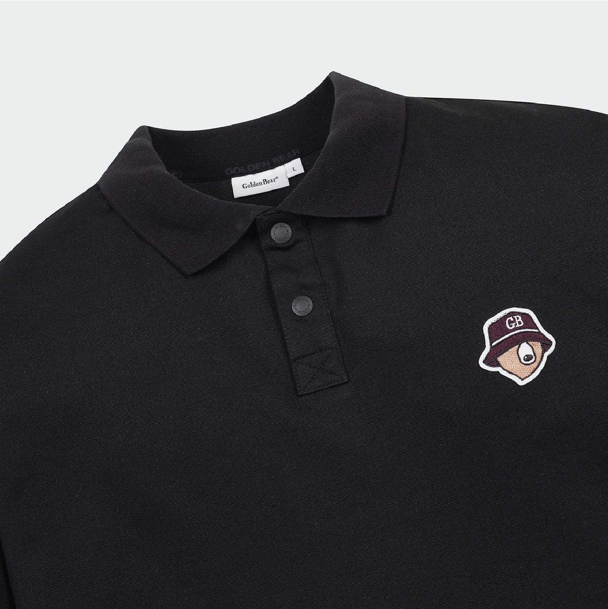 Close-up of a black polo shirt with a collar, three-button placket, and a small embroidered cartoon hat patch on the left chest. The neck label reads Schuber.