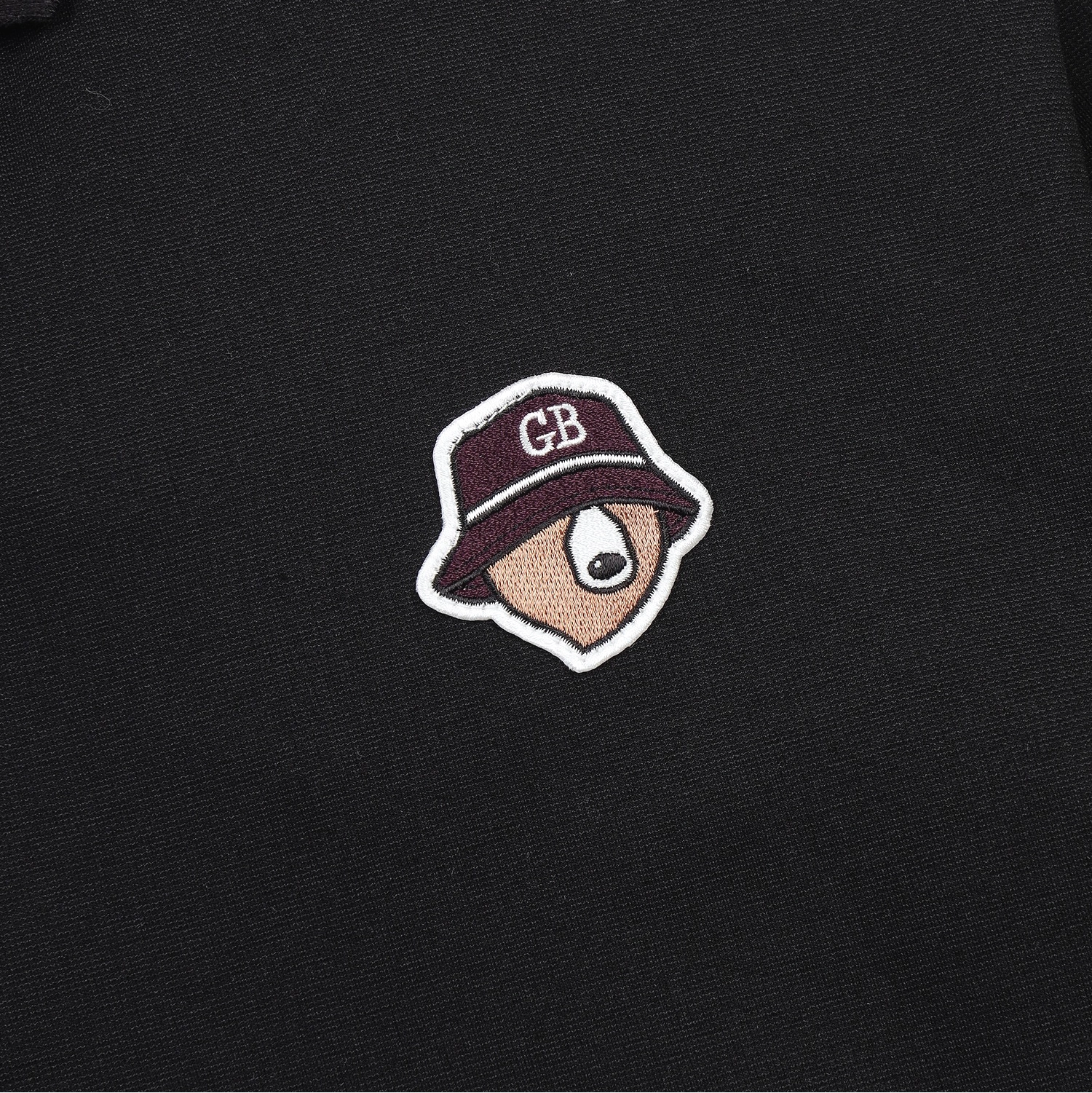A close-up of a black textured garment featuring a small embroidered patch of a cartoon face wearing a maroon bucket hat with white trim and the letters 'CB' on the hat.
