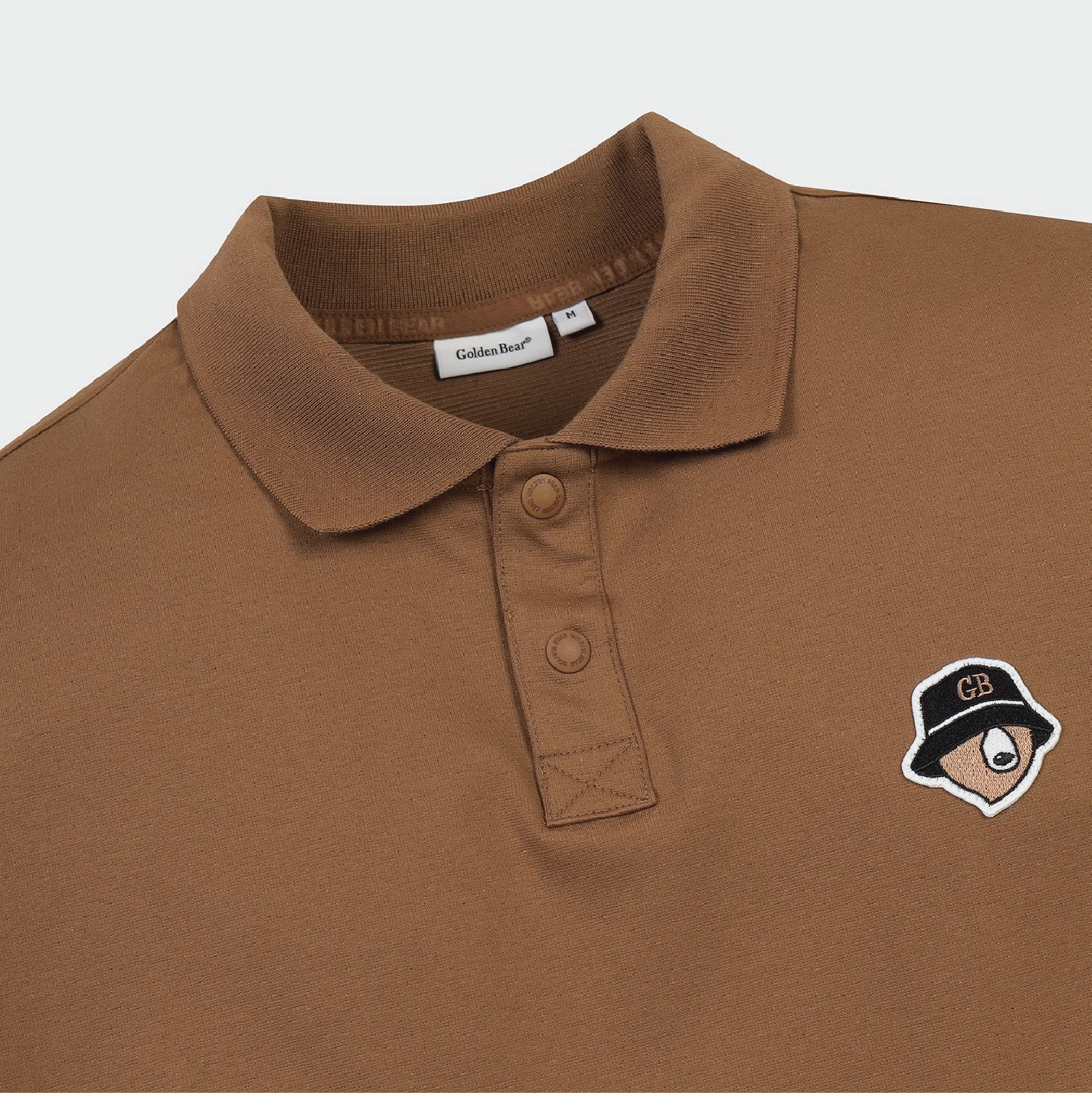 A close-up of a brown polo shirt featuring a small embroidered character patch on the left chest. The patch depicts a character wearing a black hat with a gold 'CB' logo and a white eye detail.