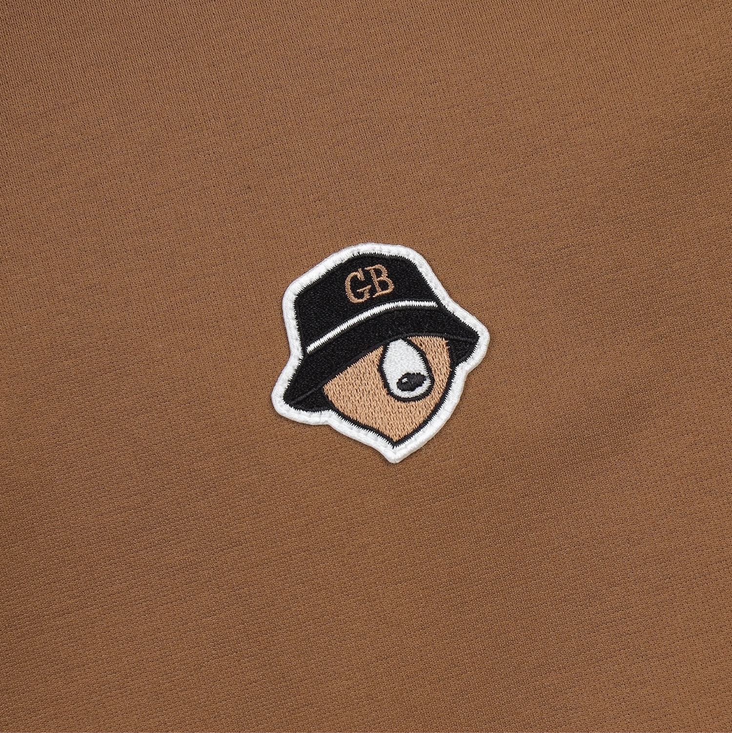 A close-up of a brown knit fabric featuring a small embroidered patch of a stylized face wearing a black bucket hat with 'GB' embroidered in gold on the front.