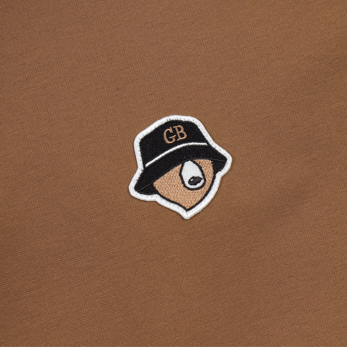 A close-up of a brown knit fabric featuring a small embroidered patch of a stylized face wearing a black bucket hat with 'GB' embroidered in gold on the front.