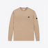 A beige long-sleeve crewneck shirt with a small embroidered patch on the left chest and a black brand patch on the right sleeve, shown against a light gray background.