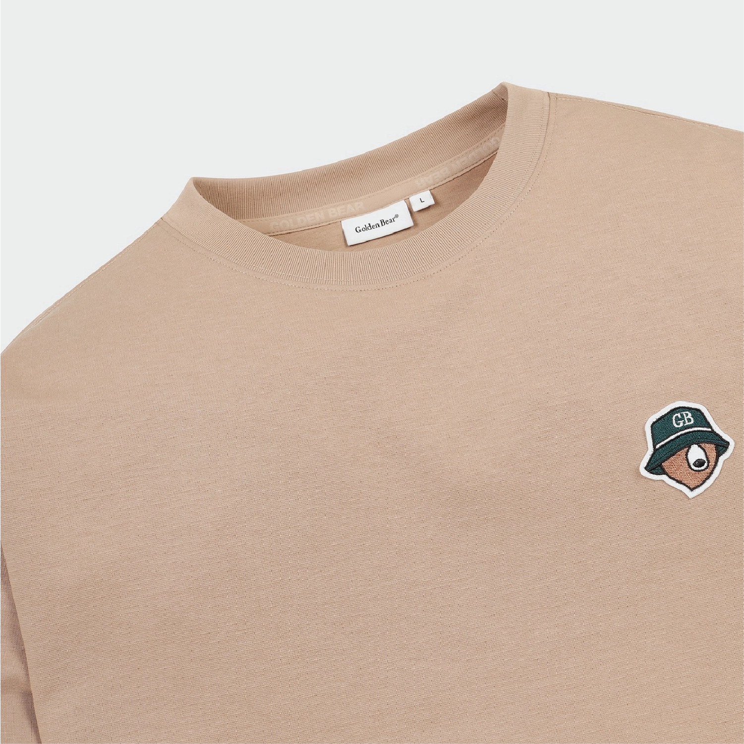 A close-up of a beige crewneck sweatshirt by GildanSoft, featuring a small embroidered cartoon bear wearing a green bucket hat with 'GB' on the chest.