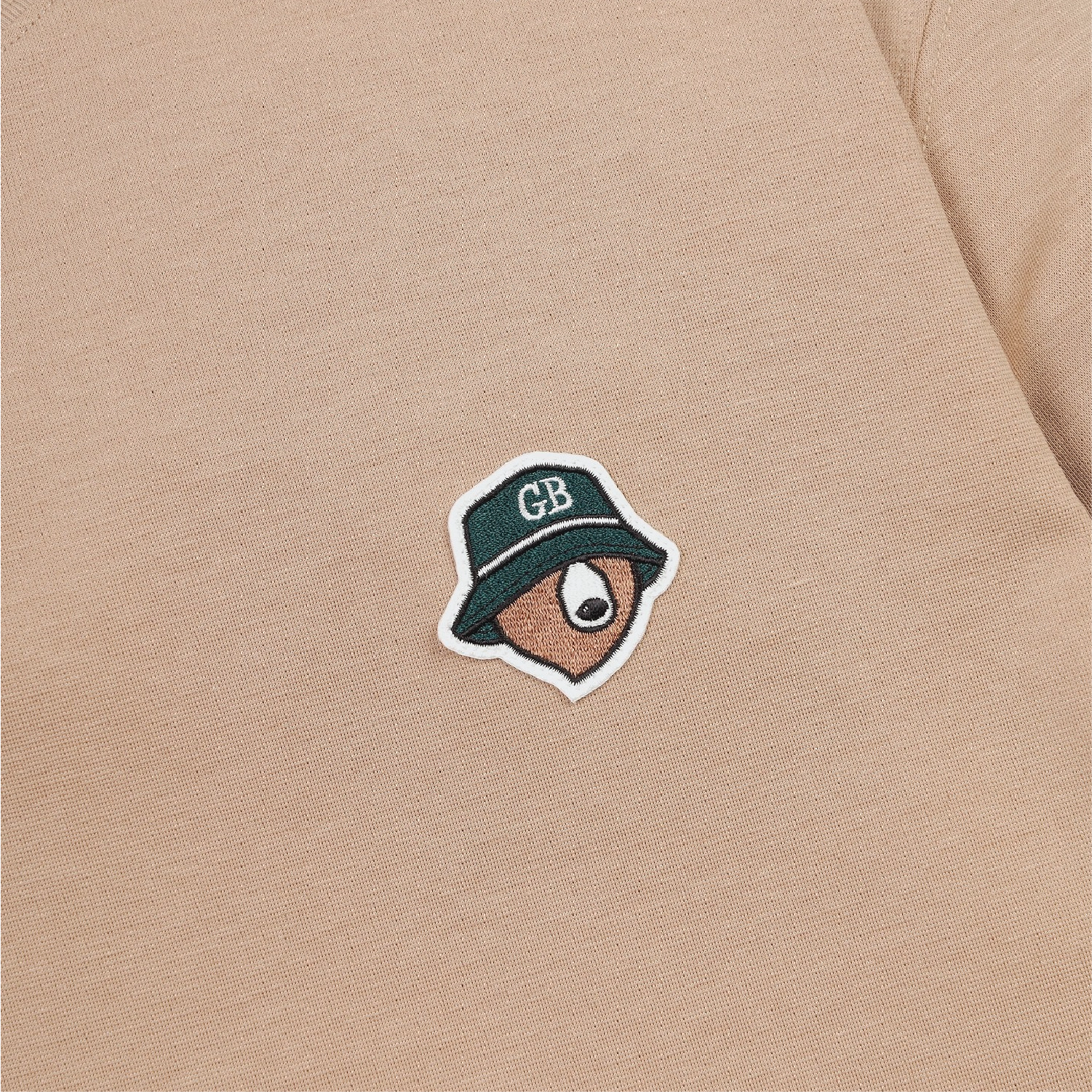 Close-up of a beige knit garment featuring a small embroidered patch of a bear wearing a dark green bucket hat with the letters 'CB' on the brim.