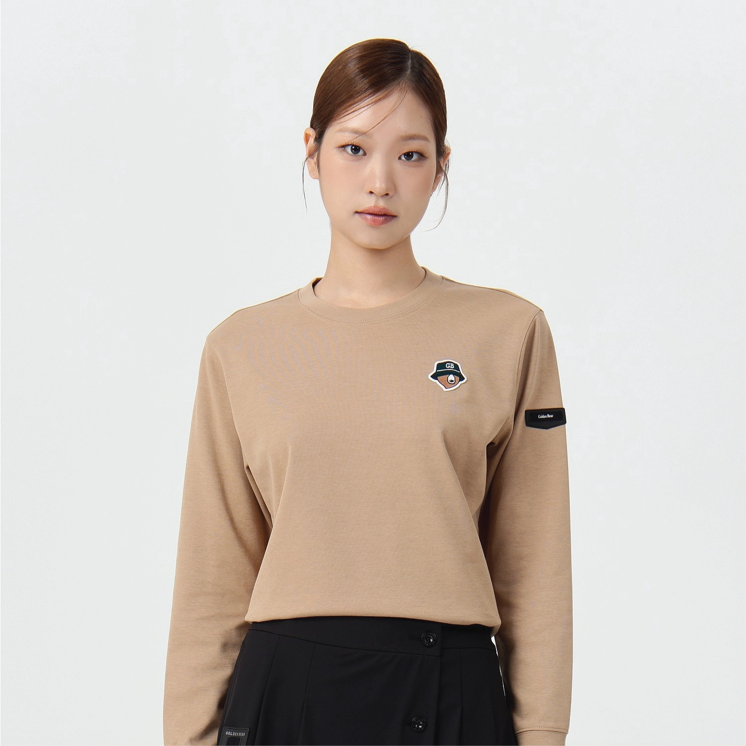 A woman modeling a beige long-sleeve crewneck sweatshirt with a small embroidered logo on the left chest and a black sleeve patch, paired with a black skirt against a plain light gray background.